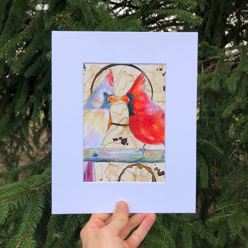 May include: A watercolor painting of two cardinals, one red and one blue, perched on a branch. The background is a coffee-stained paper with a ring around the birds.