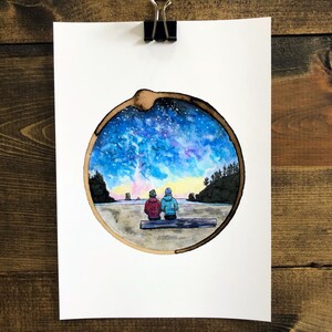 May include: Watercolor painting of two people sitting on a log, looking out at a beach scene with a starry sky above. The painting is framed in a circle, resembling a tree trunk.