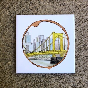 Pode incluir: Um descanso de copo de cer&acirc;mica branca com um design de anel de caf&eacute; castanho. O anel envolve uma ilustra&ccedil;&atilde;o de aquarela da ic&ocirc;nica ponte amarela de Pittsburgh, Pensilvania, com uma cidade ao fundo.