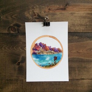 May include: Watercolor painting of a hiker with a backpack walking along a lake shore with mountains in the background. The painting is framed in a coffee ring.