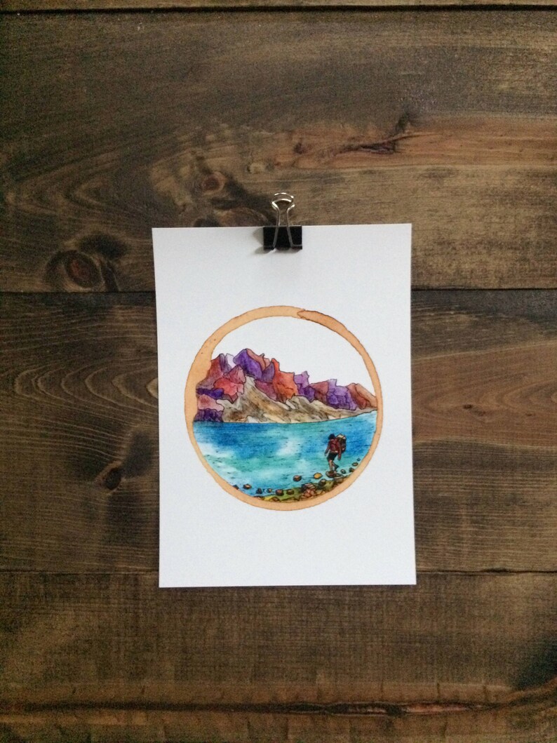 May include: Watercolor painting of a hiker with a backpack walking along a lake shore with mountains in the background. The painting is framed in a coffee ring.