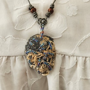 May include: A silver chain necklace with a large, round pendant. The pendant is made of a dark brown material and is covered in small, iridescent beads. The beads are arranged in a swirling pattern.