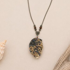 May include: A handcrafted necklace featuring a unique, irregularly shaped pendant with a mix of dark and light tones. The pendant hangs from a dark gray cord with adjustable length. The necklace is displayed on a neutral background with natural elements.