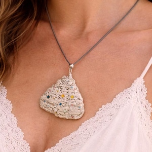 May include: A handmade pendant necklace featuring a unique, textured pendant. The pendant is a light color with colorful accents and is suspended from a gray cord. The necklace is worn against a white top.