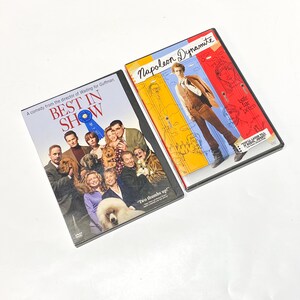 May include: Two DVD cases with movie titles "Best in Show" and "Napoleon Dynamite". The "Best in Show" case features a group of people and dogs, while the "Napoleon Dynamite" case features a cartoon-style image of a person in a vest and tie.