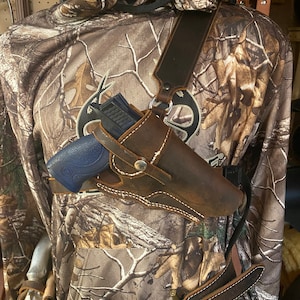 May include: A brown leather holster with a shoulder strap and a gun inside. The holster is worn on a person wearing a camouflage shirt and a brown leather belt.