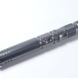 Puede incluir: Una empu&ntilde;adura de sable de luz de metal gris con un agarre texturizado y un bot&oacute;n de encendido negro.