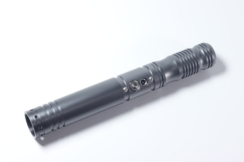 Puede incluir: Una empu&ntilde;adura de sable de luz de metal gris con un agarre texturizado y un bot&oacute;n de encendido negro.