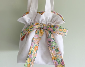 May include: A white linen tote bag with a floral ribbon bow. The bag has two white handles and a gathered top.