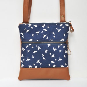 May include: A blue canvas crossbody bag with a brown leather bottom and strap. The bag has a white bird print and two zippered compartments.