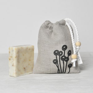 May include: A gray linen drawstring bag with a black floral design and the word "Lauren" printed on the tag. The bag is filled with a white bar of soap.