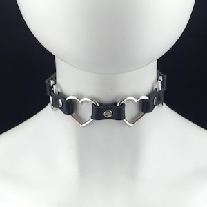 Może przedstawiać: Czarny skórzany choker z srebrnymi pierścieniami w kształcie serca. Choker jest regulowany i ma srebrną klamrę.