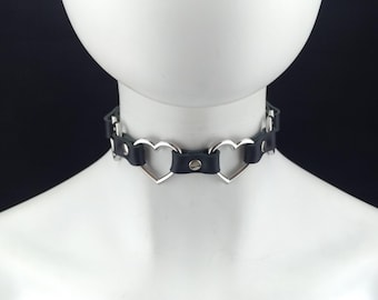 Puede incluir: Un collar de cuero negro con anillos de corazón plateados. El collar es ajustable y tiene un cierre plateado.