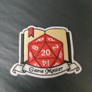 Peut inclure: Écusson brodé avec un dé à vingt faces rouge et des chiffres blancs, sur un livre ouvert. Une bannière en dessous indique "Game Master". Idéal pour les amateurs de jeux de rôle.