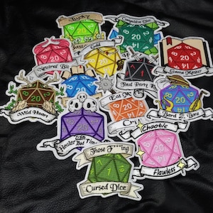 May include: A collection of embroidered patches featuring twenty-sided dice in various colors, including green, red, blue, yellow, purple, orange, and pink. Each patch includes a banner with text such as Lucky, Game Master, Wild Heart, and Cursed Dice.