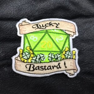 May include: Green 20-sided die with a white outline and the number 20 in the centre. The die is surrounded by four-leaf clovers and gold horseshoes. A banner above the die reads "Lucky" and a banner below reads "Bastard!"