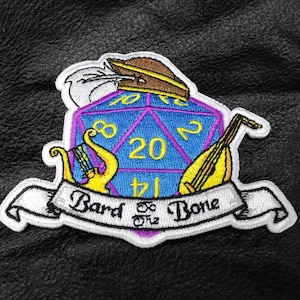 May include: Embroidered patch featuring a blue and purple twenty-sided die, a lute, and a harp. A banner reads "Bard to the Bone." A feathered hat sits atop the die. The patch is on a black leather-like surface.