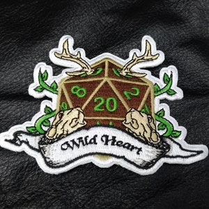 May include: Embroidered patch featuring a brown D20 die with green numbers, antlers, and two skulls. Green vines and the words "Wild Heart" on a banner complete the design. The patch is white with black and brown accents.