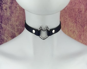Può includere: Un collare choker in pelle nera con un ciondolo a forma di cuore argentato. Il ciondolo ha due piccole orecchie di gatto sulla parte superiore.