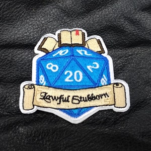 May include: A blue 20-sided die with white numbers is surrounded by scrolls and a book. The scrolls have the text "Lawful Stubborn" on them.