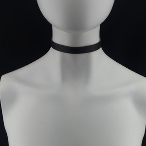 May include: A simple, solid black choker necklace worn around the neck of a white mannequin. The band is smooth, matte, and uniform in width, encircling the neck completely without any additional pendants, charms, or visible hardware closures.