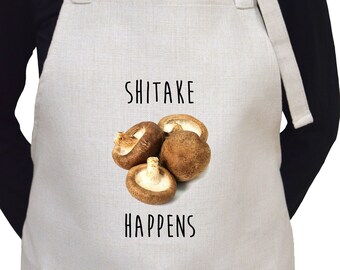 May include: A white linen apron with a pocket and adjustable straps. The apron has a brown mushroom graphic and the text "SHITAKE HAPPENS".