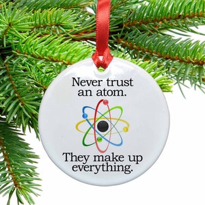 May include: White ceramic Christmas ornament with a red ribbon. The ornament features a colorful atom illustration and the text "Never trust an atom. They make up everything."
