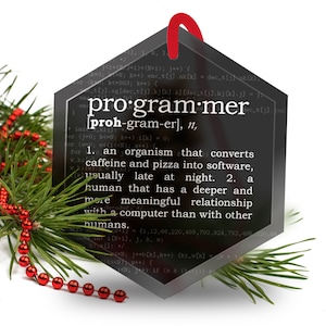 May include: A black hexagonal ornament with a white definition of the word "programmer" on a background of computer code. The ornament is decorated with a red ribbon and is sitting on a green pine branch with red beads.