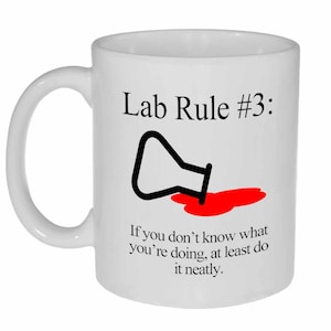 May include: White ceramic mug with a black and red graphic. The graphic depicts a beaker spilling red liquid and the text "Lab Rule #3: If you don't know what you're doing, at least do it neatly."