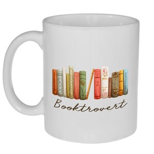 May include: White ceramic mug with a brown handle. The mug features a watercolor illustration of a stack of colorful books with the text "Booktrovert" printed below.