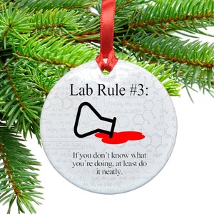 May include: A white round Christmas ornament with a red ribbon. The ornament has a black and white illustration of a beaker spilling red liquid. The text "Lab Rule #3: If you don't know what you're doing, at least do it neatly." is printed on the ornament.