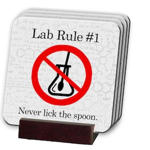 May include: A set of six white coasters with a black and white science themed background. The coasters feature a red circle with a diagonal line through it and a beaker with a spoon inside. The text "Lab Rule #1 Never lick the spoon." is printed on the coasters.