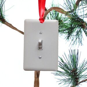 May include: A white light switch with a red ribbon hanging from the top. The switch is attached to a pine tree branch.
