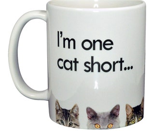 May include: White ceramic mug with the text "I'm one cat short..." in black. The mug features three cat faces peeking from the bottom. The cats have gray, brown, and yellow eyes.