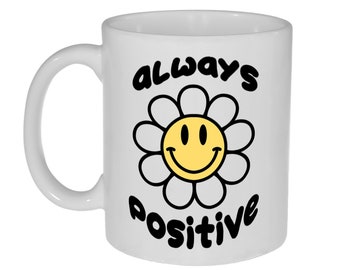 May include: White ceramic mug with a black and yellow daisy design. The daisy has a smiling face in the center and the words "ALWAYS POSITIVE" in black, bubble letters. The mug has a curved handle.