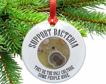 May include: White ceramic Christmas ornament with a round shape and a red ribbon. The ornament has a black and white graphic that says "Support Bacteria. They're the only culture some people have."
