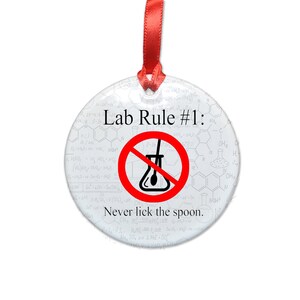 May include: A white round ornament with a red circle and a black line through it. The circle contains a beaker and a spoon. The text "Lab Rule #1: Never lick the spoon." is printed on the ornament.
