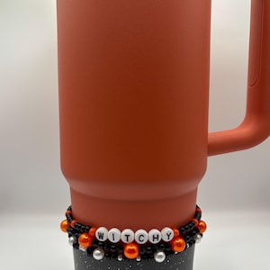 May include: A terracotta-colored Stanley tumbler with a handle, featuring a beaded accessory wrapped around its base. The accessory consists of black, orange, and white beads, with white letter beads spelling out the word WITCHY.