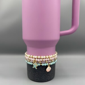 May include: A light purple Stanley mug with a black speckled base. The mug is adorned with several beaded bracelets in pastel colors, featuring charms like a starfish and seashell.