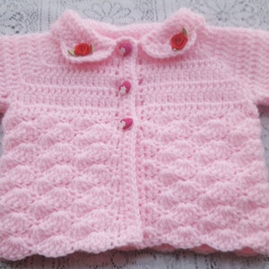 Puede incluir: Un cárdigan de bebé de color rosa tejido a crochet con cuello blanco y botones rosas. El cárdigan tiene un patrón de punto decorativo y está adornado con dos pequeñas flores rojas.