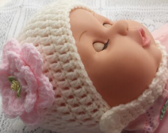 May include: A white crocheted baby hat with a pink flower on the side. The hat has a tie that goes under the chin.