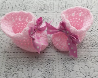 Op de afbeelding: Een paar handgemaakte, lichtroze gehaakte babyschoentjes. Elk schoentje heeft een geschulpte rand en een roze lint met witte stippen dat tot een strik is geknoopt. De schoentjes zijn ontworpen voor baby's.
