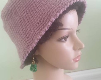 Può includere: Un cappello a secchiello rosa lavorato a uncinetto con un orecchino a nappa verde. Il cappello è indossato da un manichino con una collana di perline verde acqua e rosa.