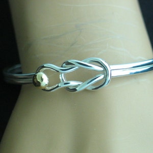 May include: A silver bangle bracelet with a knot design and a small gold bead accent.