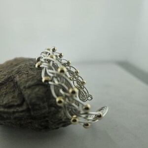 May include: A cuff bracelet constructed from multiple intertwined silver-toned wires, featuring small, evenly spaced gold-toned spherical beads integrated throughout the woven metal structure, creating a textured and layered appearance.