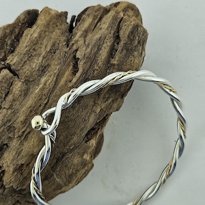 May include: A silver and gold-tone twisted bangle bracelet. The bracelet features a braided design with silver and gold-colored metal strands. A small gold-colored ball is at the clasp. The bracelet is displayed on a piece of textured, brown wood.