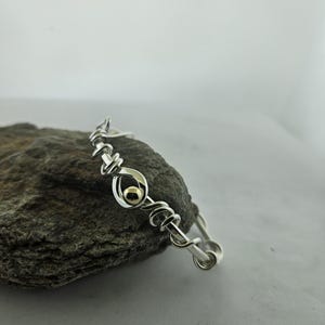 May include: A silver wire-wrapped bangle bracelet featuring a central loop design with a small, polished gold-tone bead nestled within the center. The bracelet is constructed from a continuous silver wire coiled into a series of decorative loops.