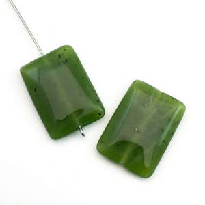 Op de afbeelding: Twee rechthoekige, doorschijnende, olijfgroene jade kralen. Eén kraal is geregen aan een zilverkleurige draad. De kralen hebben afgeronde hoeken en een gepolijst oppervlak. De jade heeft donkere insluitsels en een lichte kleurvariatie.