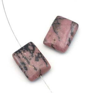 Peut inclure: Deux perles rectangulaires en rhodonite, rose avec des veines noires. Elles sont enfilées sur un fil fin argenté, sur fond blanc. Les perles polies ont des bords arrondis, parfaites pour la création de bijoux.