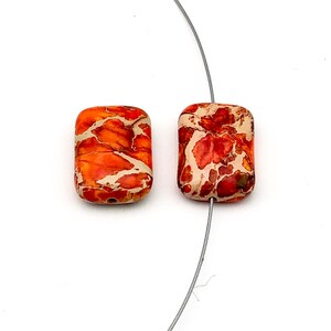 Peut inclure: Deux perles rectangulaires aux bords arrondis. Les perles sont orange avec des veines crème et marron. Un fin fil gris traverse le centre de chaque perle. Les perles sont posées sur un fond blanc.